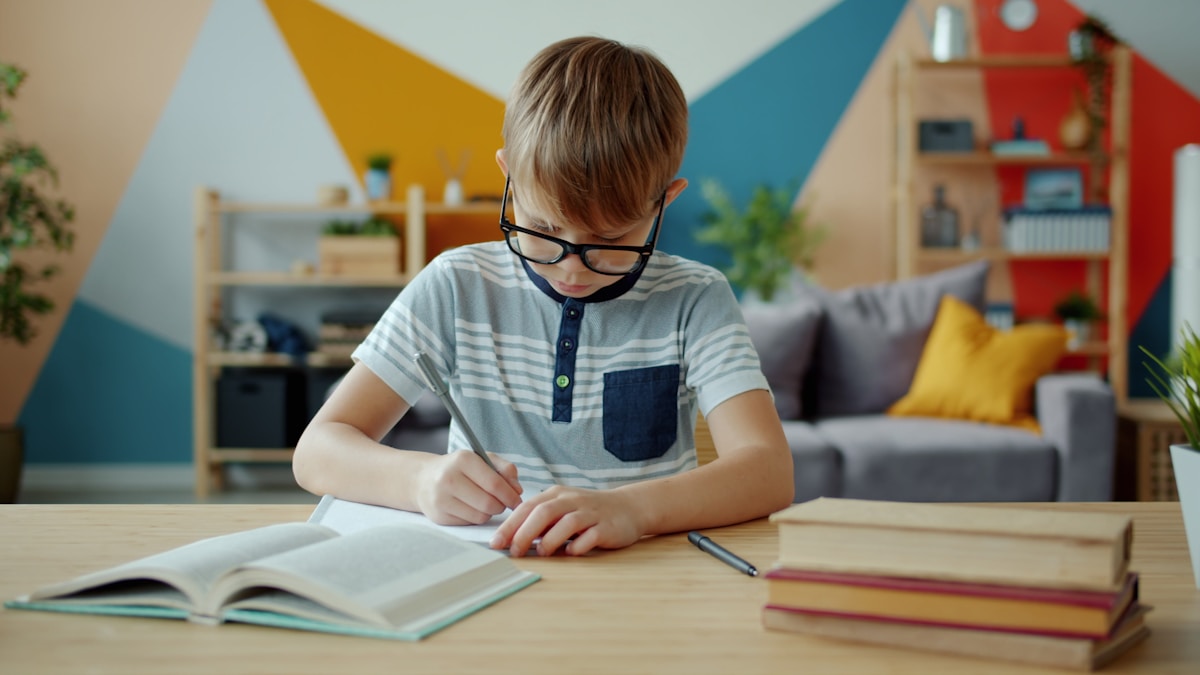 A young child wearing glasses, concentrating as they write at a desk surrounded by books