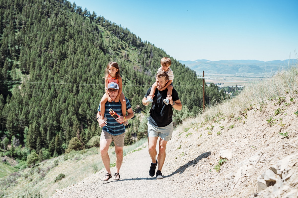 Two parents carrying young children on their shoulders along an outdoor trail