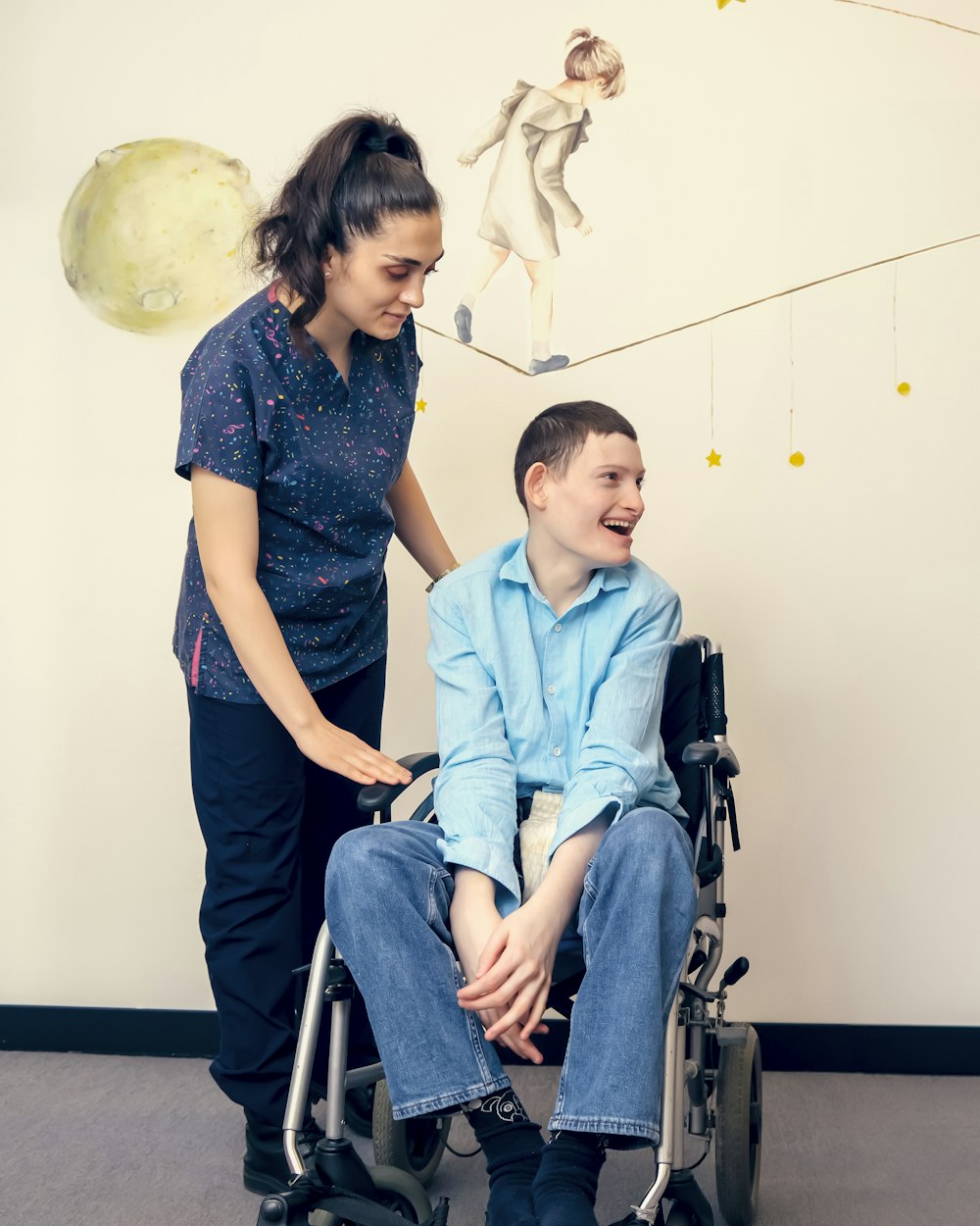 A support worker beside a young man who uses a wheelchair, both smiling, in a calm assessment room